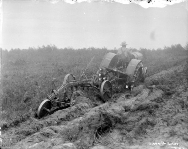 Three-quarter view from right rear of a man driving a 10-20 tractor for pulling a plow to break up a field.