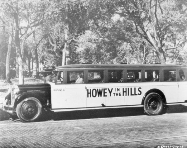 Left side profile view of Bus No. 4, with a sign on the side reading: "Howey in the Hills." A man is sitting in the driver's seat. The five rows of seats in the back of the bus are empty of people.