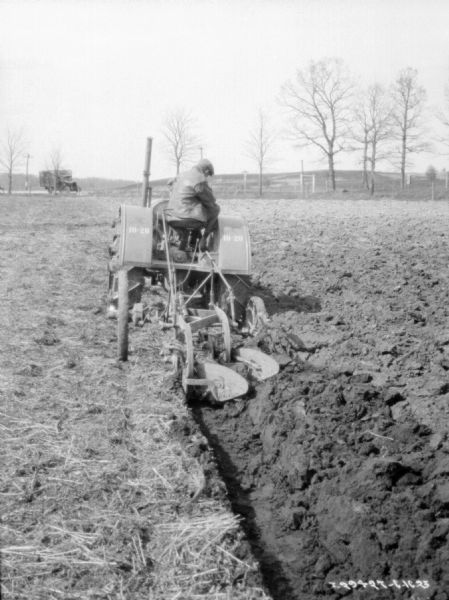 Rear view of a man driving a 10-20 tractor to pull a flow in a field. A truck is parked in the background.