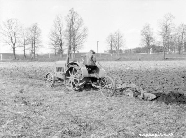 Three-quarter view from left rear of a man using a 10-20 tractor to pull a plow in a field.