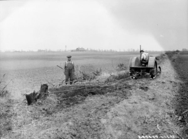 One man is standing and watching while a man driving a tractor is pulling a stump from a field. There are farm buildings in the background.