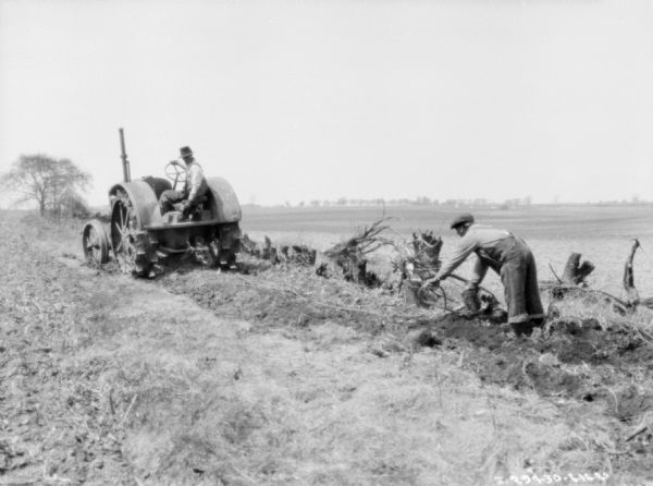 Three-quarter view from left rear of a man driving a 10-20 tractor pulling a plow in rough ground. A man on the right is helping with a rope being used to pull stumps from the field.