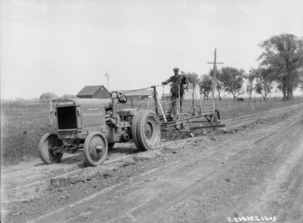 Three-quarter view from front left of a man driving an industrial tractor pulling a road grader. In the background is a field, and farm buildings and large stacks of hay are in the distance.
