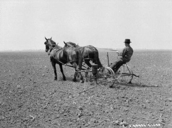 Three-quarter view from left rear of a man on a horse-drawn planter in a field.