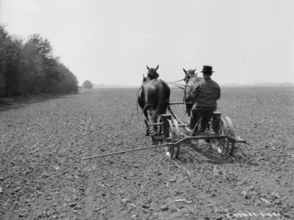 Rear view of a man using a horse-drawn planter in a field. Trees line the field on the left.