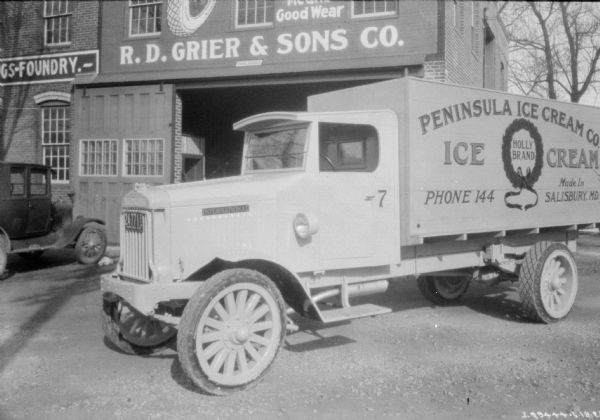A dairy delivery truck parked in front of a building with a sign that reads: "R.D. Grier & Sons Co." The sign painted on the side of the truck reads: "Peninsula Ice Cream Co."