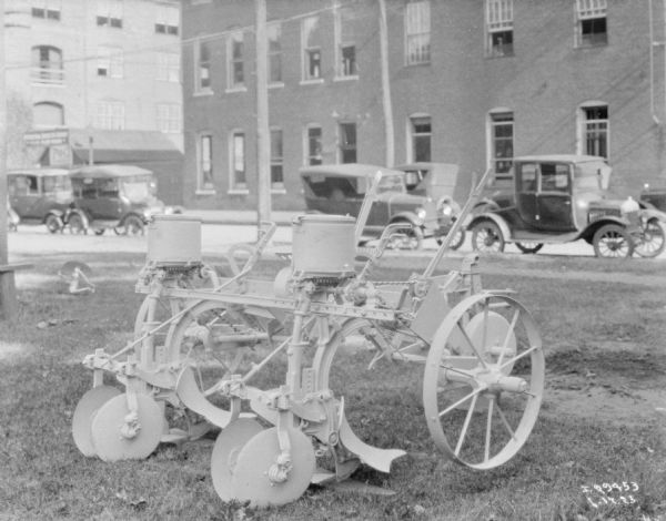 Corn planter on display on a lawn outdoors. In the background, automobiles are parked along a street. There are brick buildings across the street in the background.