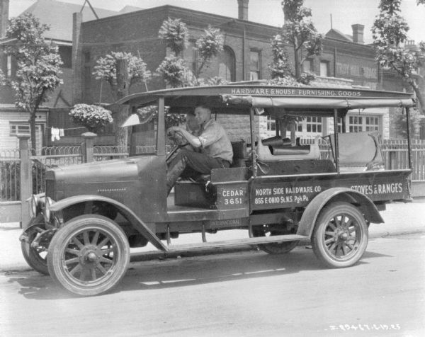 Man driving a hardware store delivery truck on a street. The sides of the truck are open, with the covers rolled up near the roof. There is a man in the passenger seat. Painted on the side of the truck are signs that read, in part: "North Side Hardware Co., 855 E-Ohio St. N.S. Pgh. Pa."