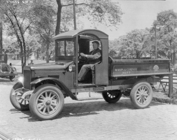 Man sitting in a delivery truck near a park. There is no driver's side door, but there is a rolled-up cloth at the top of the door. The truck bed is open. The painted sign on the side of the truck reads: "Morris & Company."