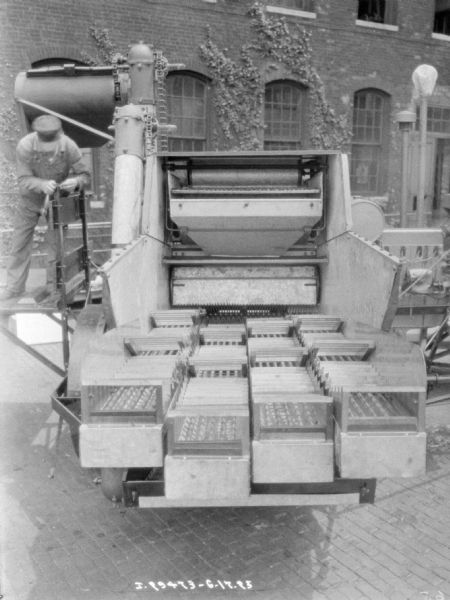 Rear view of a harvester thresher parked on cobblestones near a brick building. A man is standing on the side of the thresher on the left.