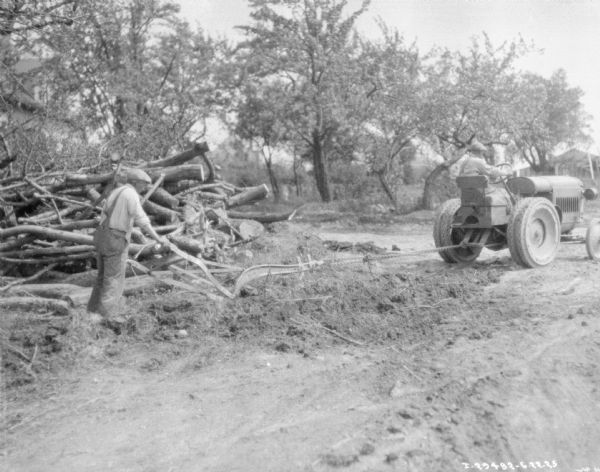 Right side view of a man standing behind a walking plow being pulled by a man driving a tractor.