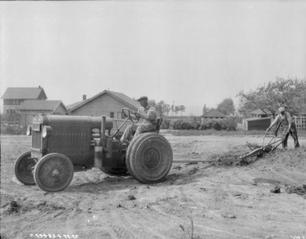 Left side view of a man standing behind a walking plow being pulled by a man driving a tractor. There are farm buildings in the background.