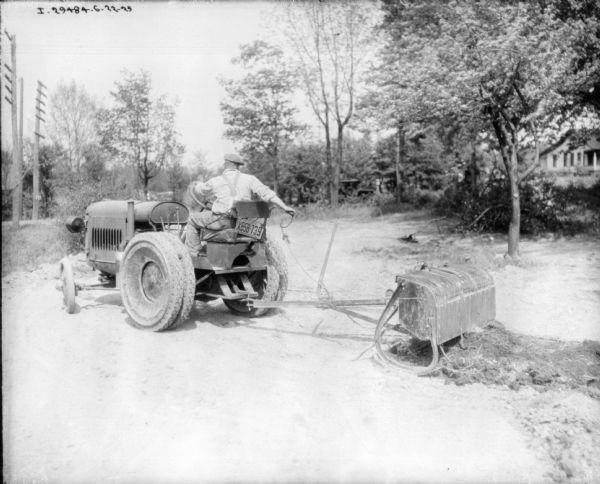 Three-quarter view from rear left side of a man driving a tractor to pull a piece of equipment. The man is pulling a chain that appears to be dumping material onto the ground. There is a commercial license plate on the back of the seat of the tractor.