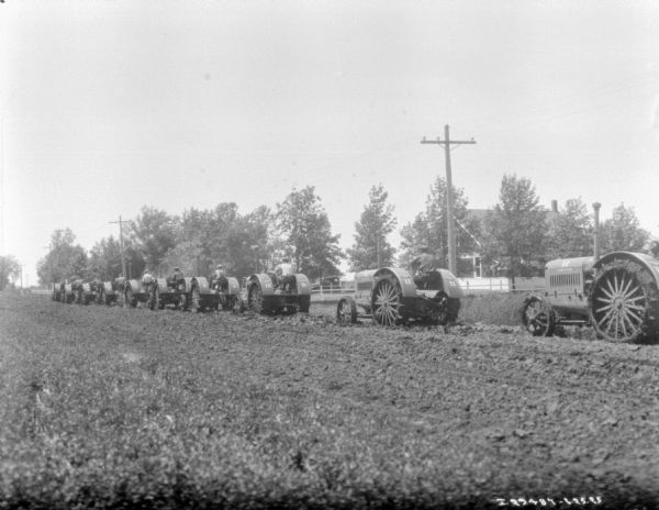 View across field towards a group of men driving kerosene tractors in a long row down a field.