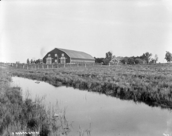 View across a stream in a field towards a large barn. There is a farmhouse in the background.