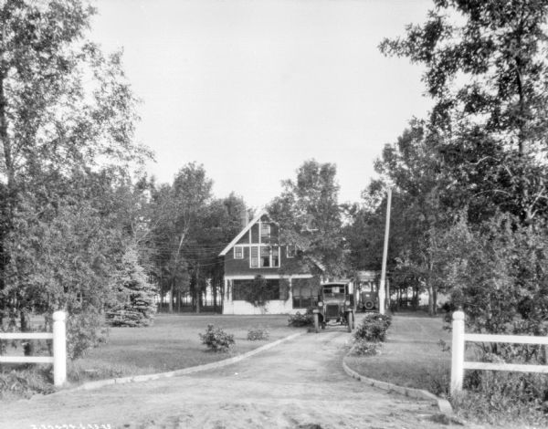 View from road of trucks parked in the driveway next to a farmhouse. One of the trucks or an automobile is parked under the coach gate or carriage porch on the right of the farmhouse.