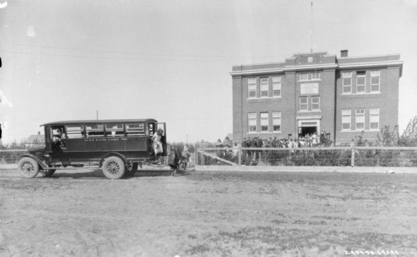 Children are getting off a school bus which is parked near the gate of a fence surrounding a brick school building. The children are exiting the bus through the back door, which has a set of steps.