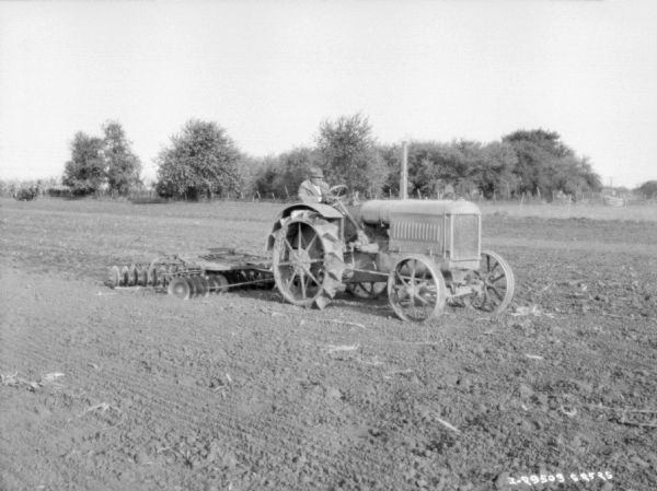 Three-quarter view from front right driving a tractor pulling a disk harrow.