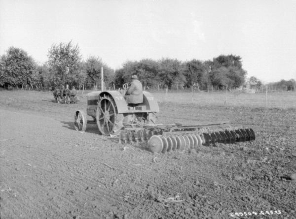 Three-quarter view from left rear of a man on a tractor pulling a disk harrow. In the background is a man on a horse-drawn agricultural implement.