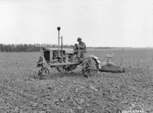 Three-quarter view from front left of a man driving a Farmall tractor to pull a disk harrow.