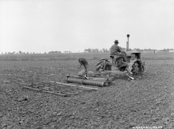Three-quarter view from right rear of a man driving a tractor pulling a disk harrow in a field. There are farm buildings in the background.