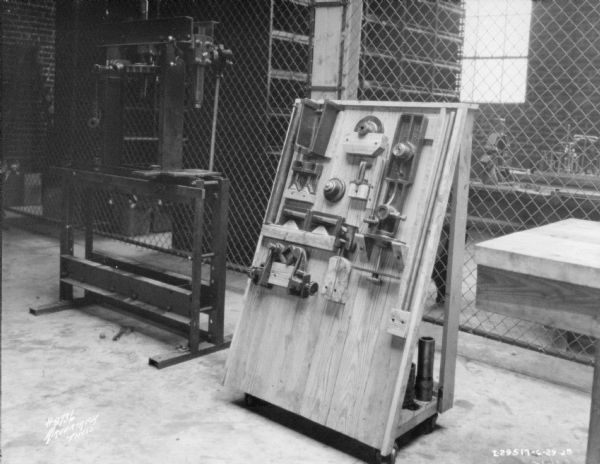View of repair shop, with a piece of machinery on the left, and a vertical wood stand on wheels that is holding parts. In the background is a wall made of wire fencing that separates the shop from another room. 
