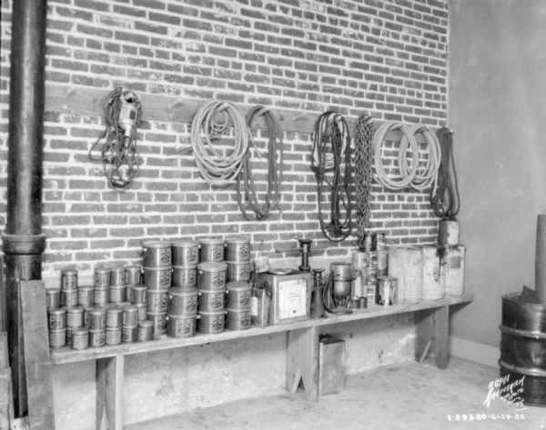 View of bench and rack against a brick wall. The bench is stacked with cans of grease and other supplies. The wood rack is holding hoses, ropes, chains, etc.