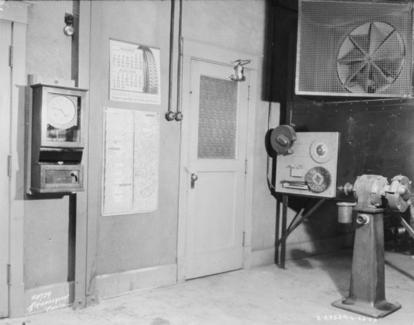 Interior view of a repair shop at a dealership. There is a clock on the wall to the left of a calendar and a door. On the right is a piece of machinery, machinery parts on a board on a wall. Near the ceiling is a large fan.