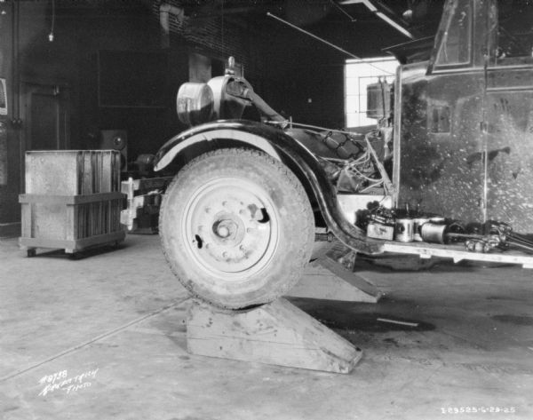 Interior view of a repair shop. In the foreground is the front of a truck up on blocks. In the background is a cart loaded with materials.