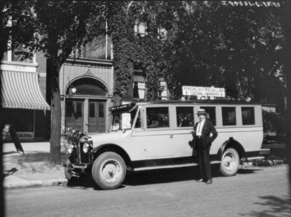 View across street towards a man posing in front of a bus parked at the curb. A sign on top of the bus reads: "International Chassis Motor Coach Ltd. Body, Chatham Products." In the background are buildings, one of which has a sign for the YMCA.