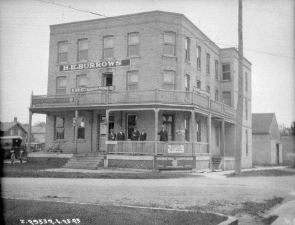 View across street towards the H.E. Burrows dealership, a large, three-story brick building. A group of men are standing on the porch. 