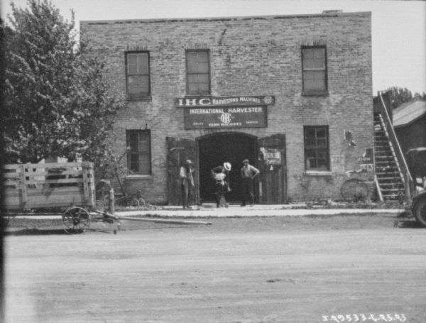 View across road towards three men on the sidewalk inspecting a cream separator in front of a dealership. Behind them are two open doors of a large arched entrance to the dealership, which is a two-story brick building. The doors are open, and a sign above reads: "IHC Harvesting Machines." A wagon is parked on the left.