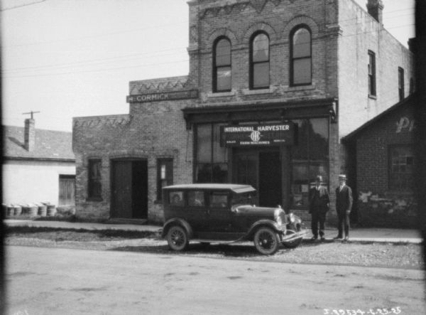 View across street towards two men posing on the sidewalk with a touring car in front of a dealership. Two people are sitting in the back seat of the car.
