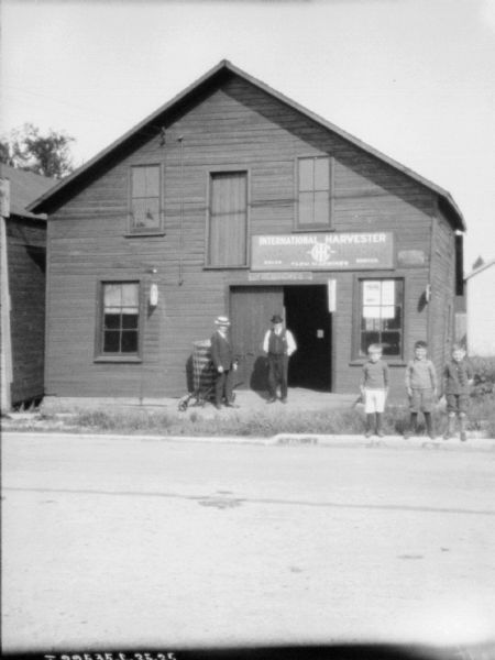 View across street towards two men and three boys posing on the sidewalk in front of an International Harvester dealership.