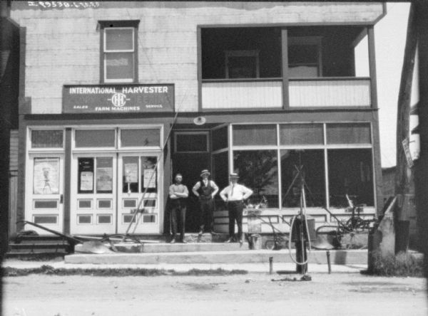 View across street towards men posing in front of an International Harvester dealership. Agricultural implements are set up on the steps, and there is a sign for "Free Air" on a post on the right.