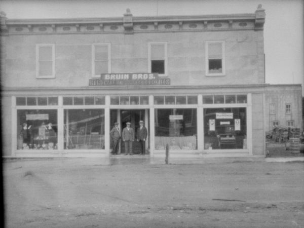 View across street towards three men posing infront of a dealership. The sign above the entrance reads: "Bruin Bros. Hardware and Accessories."