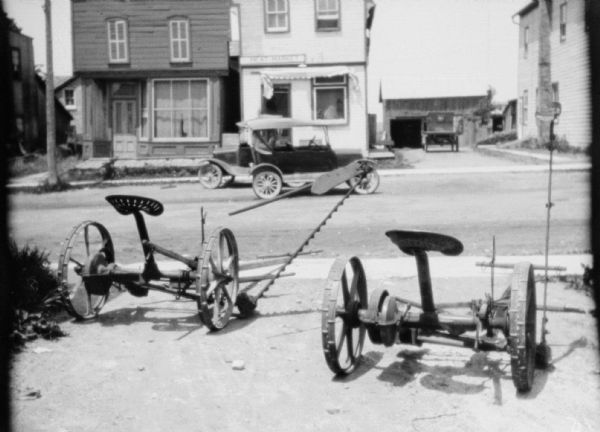 View of mowers parked on the pavement near the street. There are two automobiles in the street. Buildings and garages are in the background. One of the buildings has a sign above the entrance that reads: "Meat Market."
