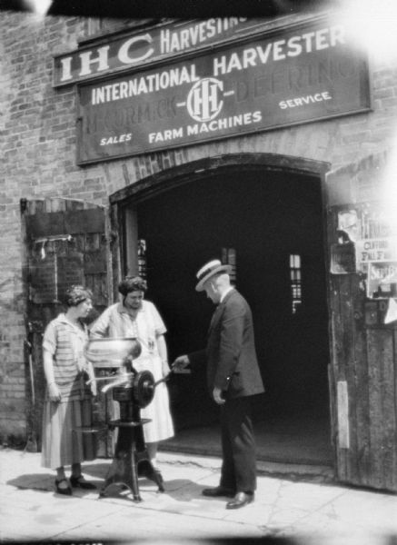 Two women are listening to a man while standing on the sidewalk in front of an International Harvester dealership. They are looking at a cream separator in the large open doorway of the dealership.