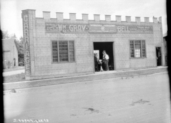 View across street towards three men standing in the open doorway of a dealership. The men are looking at a cream separator. The building is a one-story building, and a sign at the corner reads: "Carriages" and another on the side reads: "Wm. Gray-" but is unreadable because a sign for International Harvester has been placed over it.