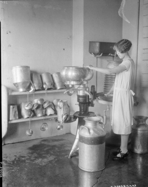 View of a woman working in a room at a dairy with a McCormick-Deering Primrose cream separator. There are metal milk cans on shelves behind her.