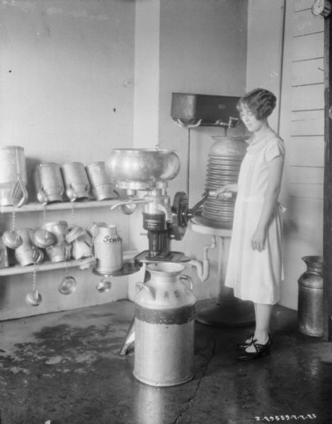View of a woman working in a room at a dairy with a McCormick-Deering Primrose cream separator. There are metal milk cans on shelves behind her.