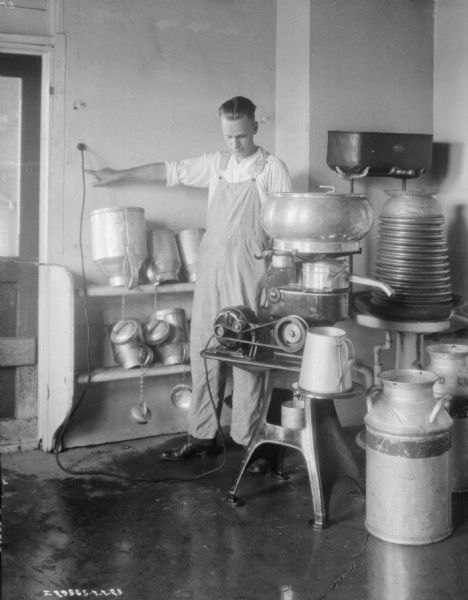 View of a man working in a room at a dairy with a McCormick-Deering Primrose cream separator. There are metal milk cans on shelves behind him.