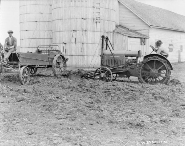 Two men in a barnyard. The man on the left is sitting on a horse-drawn manure spreader, and the man on the right is on a tractor with a front loader. There are two silos and a barn in the background.