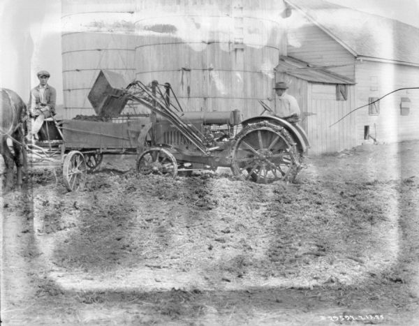 A man on the right is operating a tractor with a front loader to fill up a manure spreader. A man is sitting on the horse-drawn manure spreader. There are two silos and a barn in the background.