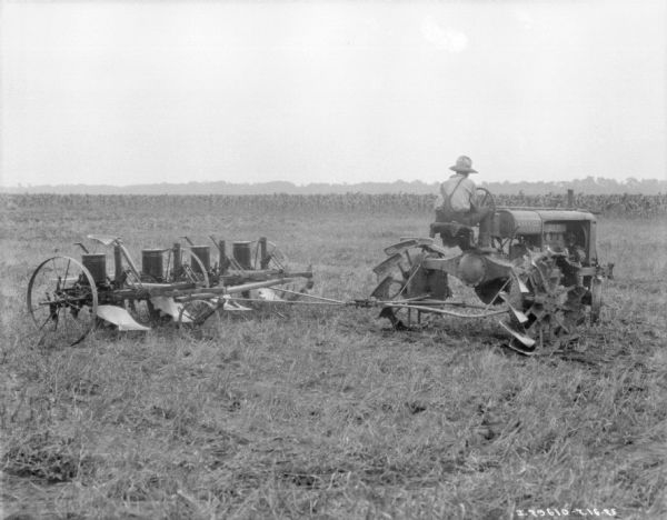 Three-quarter view from right rear of a man using a Farmall tractor to plant in a field.
