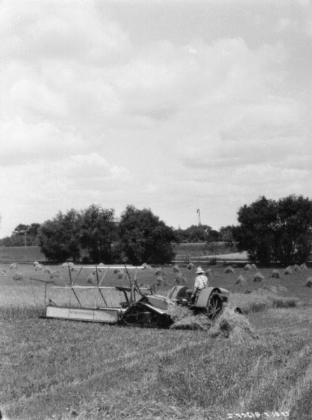 View across field towards a man using a tractor to pull a binder. There are piles of harvested grain in the field, and a windmill is in the distance.