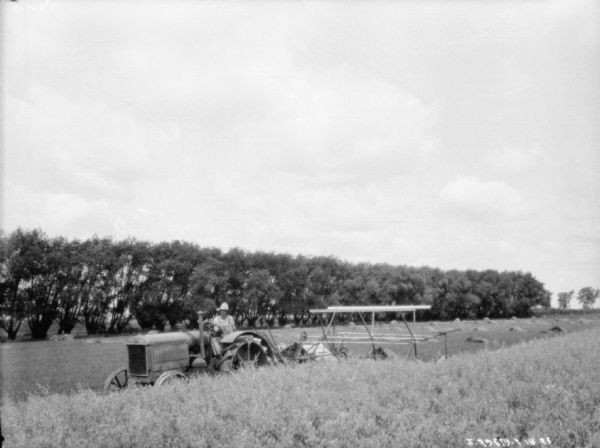 View across unharvested section of field towards a man driving a tractor pulling a binder. Piles of harvested grain are in the background.