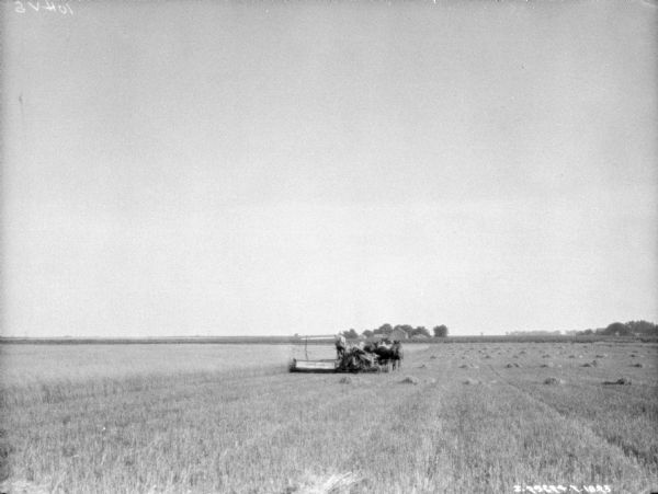 View down field towards a man using a horse-drawn binder in a field. Farm buildings are in the far background.