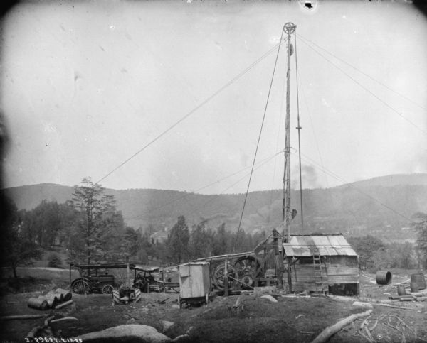 View down slope towards a tractor powering a sawmill in a forest. There is a valley and hills in the background.