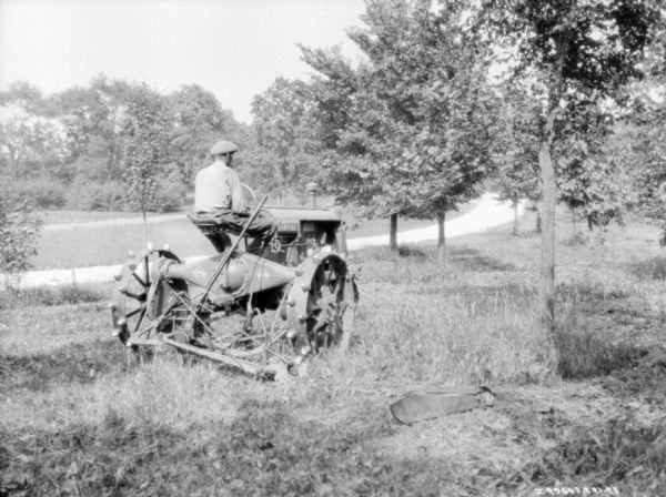 Three-quarter view from right rear of a man using a Farmall tractor to pull a mower to mow a field.